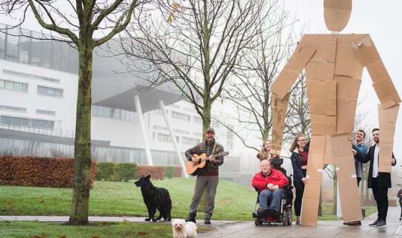 Students outside Ӱֱholding a giant card board person up whilst a man with a dog plays guitar