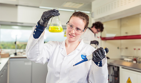 A Ӱֱstudent wearing PPE and holding up a beaker of yellow liquid